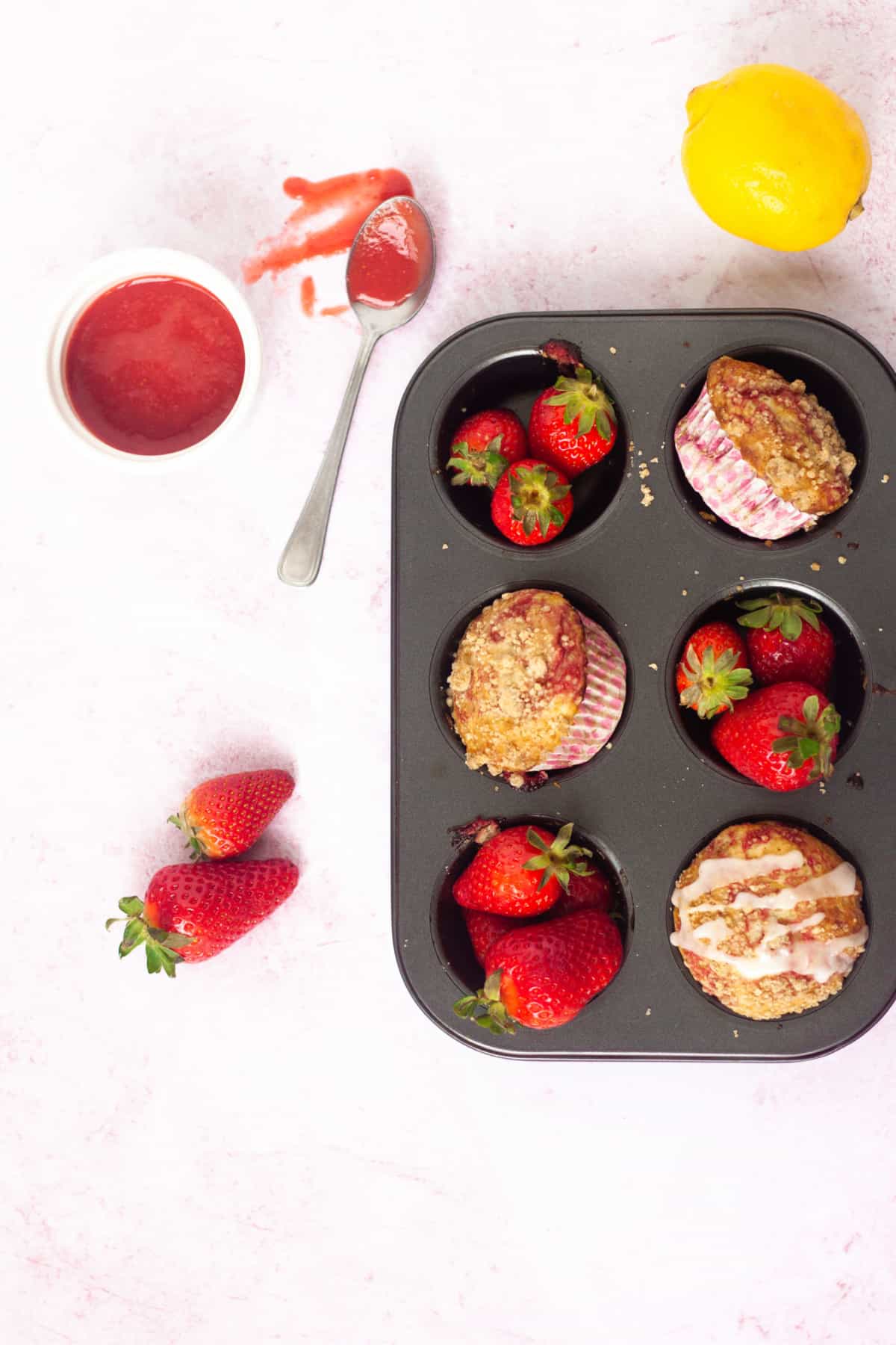 Eggless Strawberry Swirl Muffins in muffin tray surrounded by strawberry puree, fresh strawberries, and a lemon