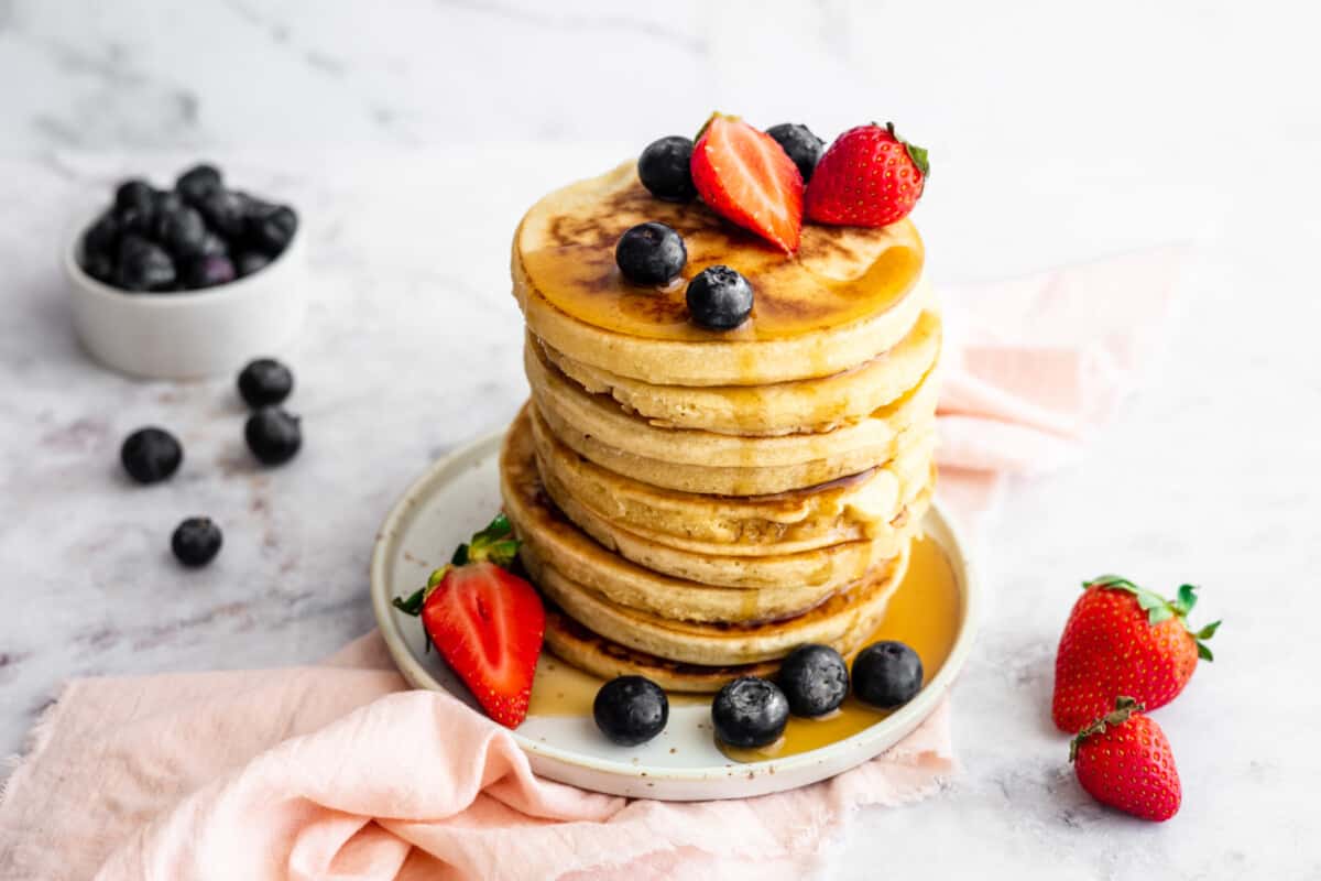 A stack of fluffy almond milk pancakes with maple syrup, strawberries and blueberries.